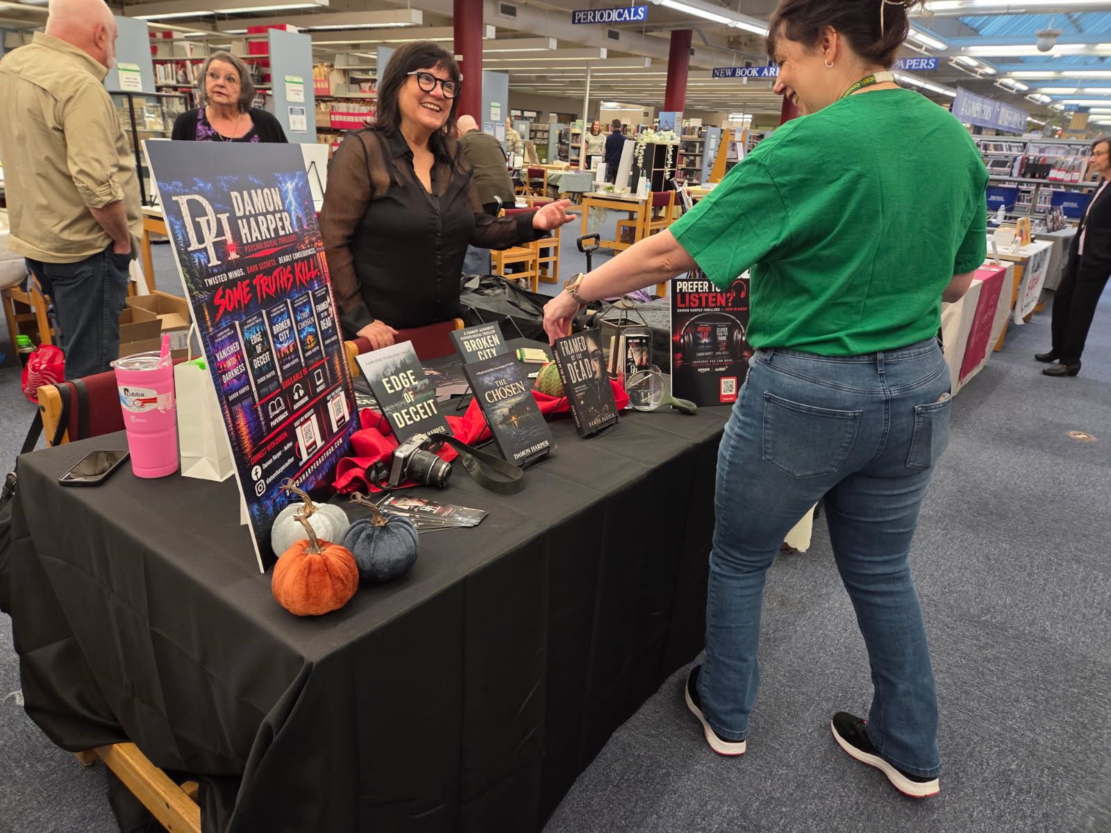 A Damon Harper table at the library book festival: a tall banner stand, paperbacks spread across a black tablecloth, autumn pumpkins, and a visitor in a green shirt browsing while a woman smiles behind the table.